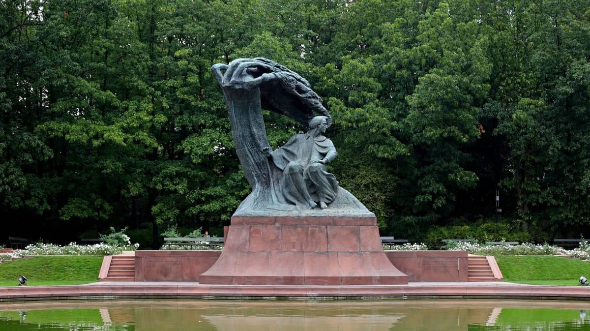 Duży pomnik Chopina pod stylizowanym drzewem w parku, z oczkiem wodnym i zielonymi drzewami w tle w Warszawie.