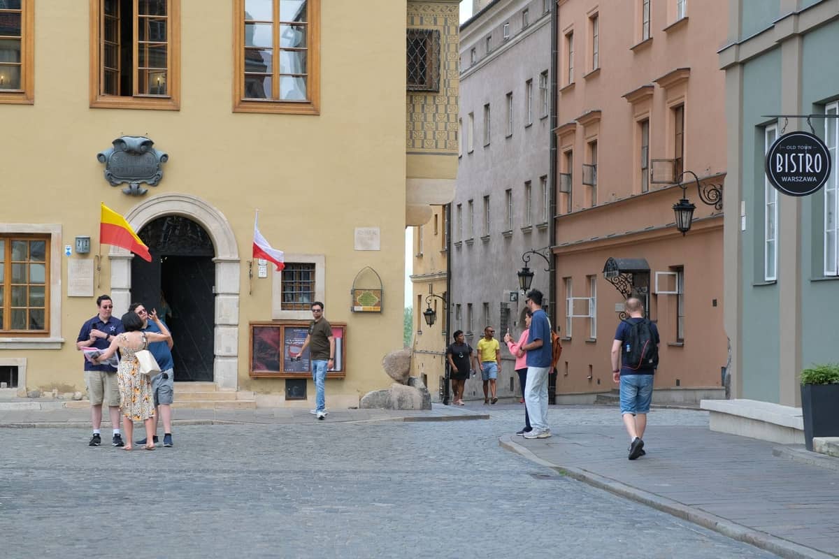 Ludzie spacerujący i rozmawiający na brukowanej ulicy otoczonej kolorowymi zabytkowymi kamienicami i szyldem bistro w Starym Mieście w Warszawie.