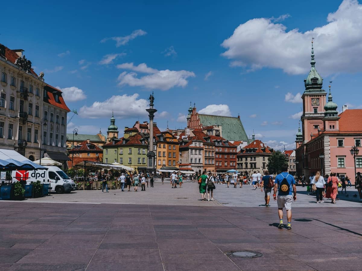 Słoneczna letnia scena Starego Miasta w Warszawie i Placu Zamkowego z przechadzającymi się ludźmi, kolorowymi historycznymi budynkami i błękitnym niebem z chmurami.