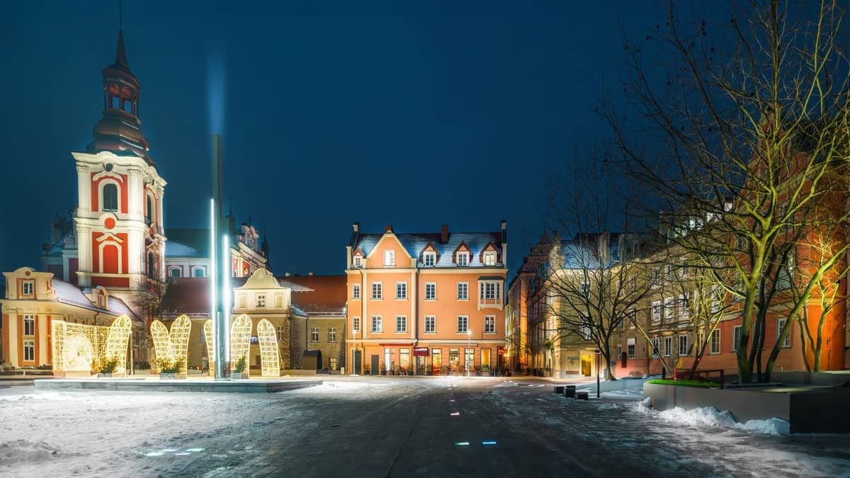 Alun-alun Kota Tua Warsawa pada malam hari dengan salju di tanah, bangunan bersejarah yang diterangi cahaya, dan menara gereja di bawah langit musim dingin yang gelap.