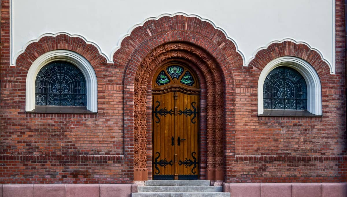 Primer plano de la ornamentada entrada de ladrillo con arcos de la Sinagoga Nożyk en Varsovia, con ventanas decorativas y una puerta de madera.
