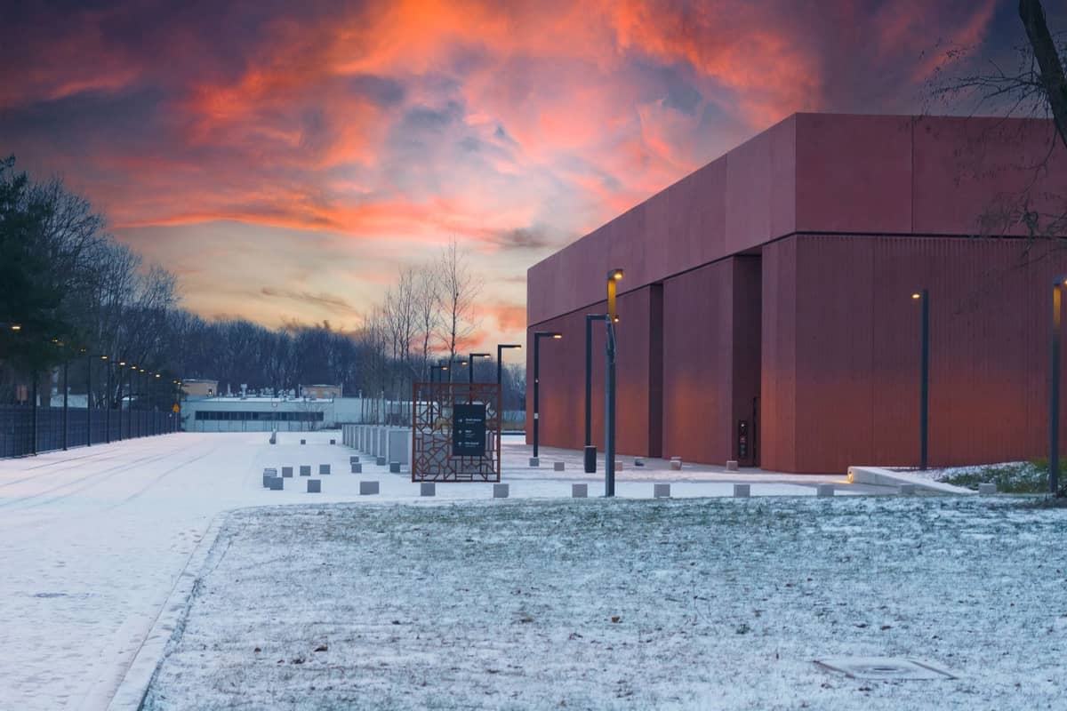 Edificio moderno con arquitectura minimalista y fachada rojiza, fotografiado al atardecer con nieve en el suelo y árboles al fondo.