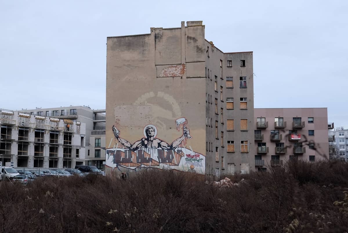 Solar vacío con arbustos crecidos, un aparcamiento y un edificio de apartamentos de Varsovia con grandes grafitis en la pared—una escena urbana que evoca huellas del pasado.