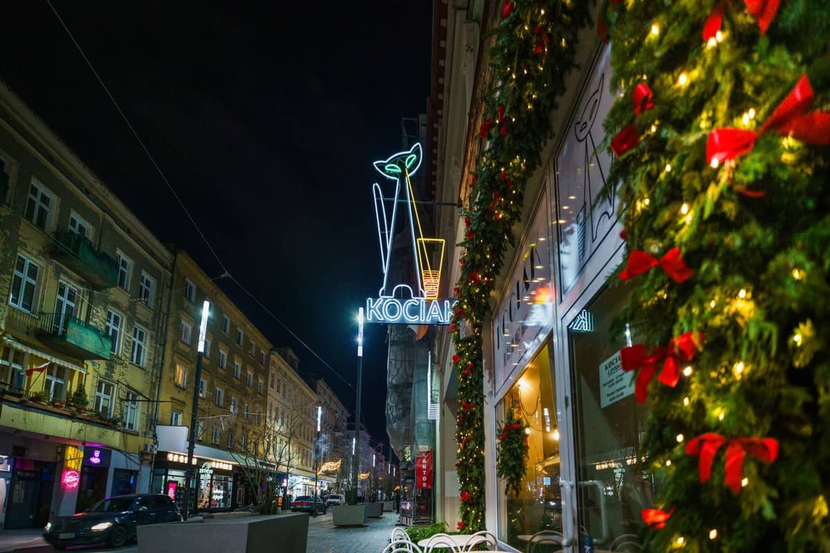 Vista notturna di una strada di Varsavia con decorazioni festive, insegna al neon di un bar e classici edifici di appartamenti lungo il marciapiede.