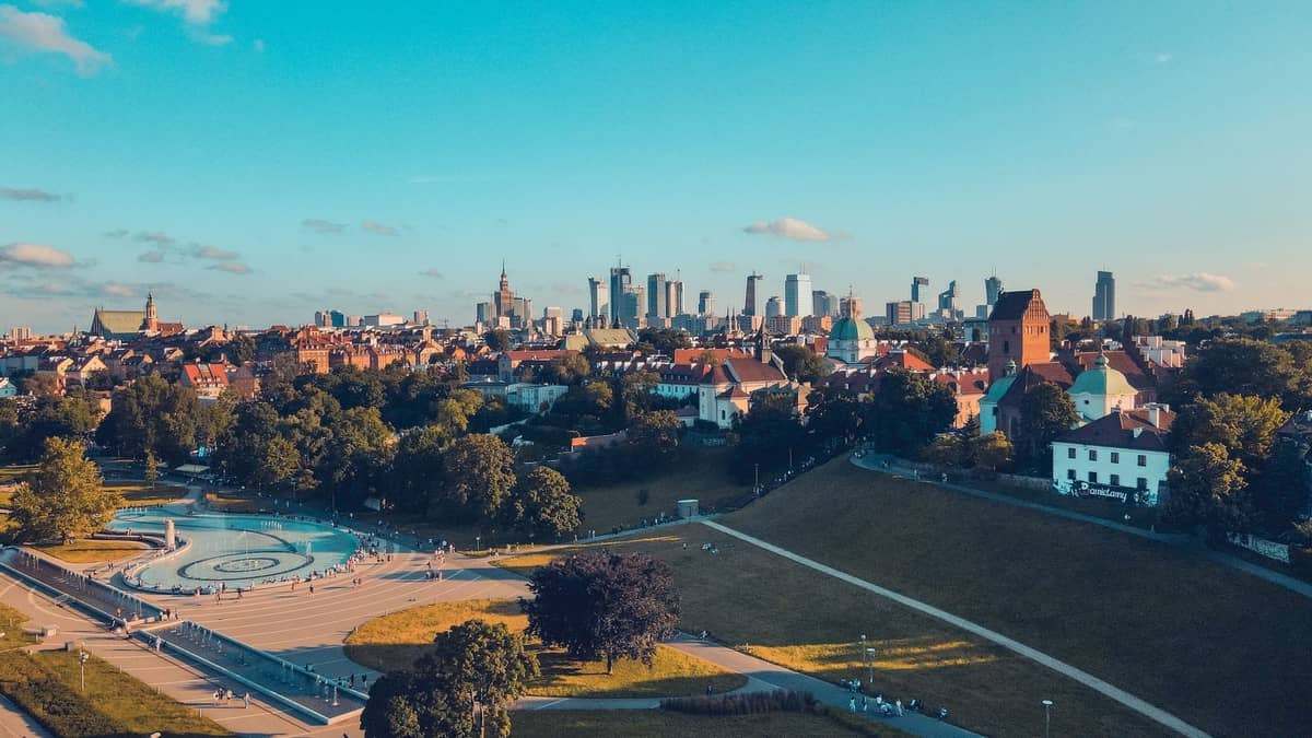 Vista aérea de un amplio parque con una fuente circular, edificios históricos de la ciudad y el horizonte de Varsovia al fondo.