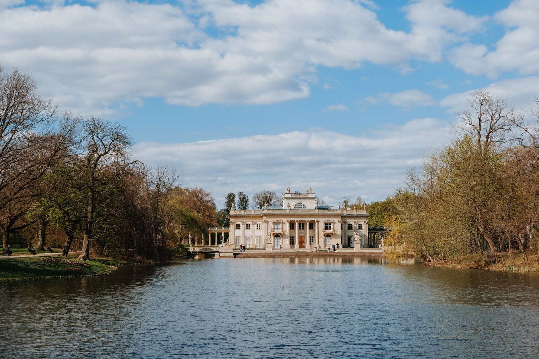 Palácio na Ilha no Parque Łazienki de Varsóvia, cercado de árvores e refletido em um lago tranquilo sob um céu azul com nuvens esparsas.