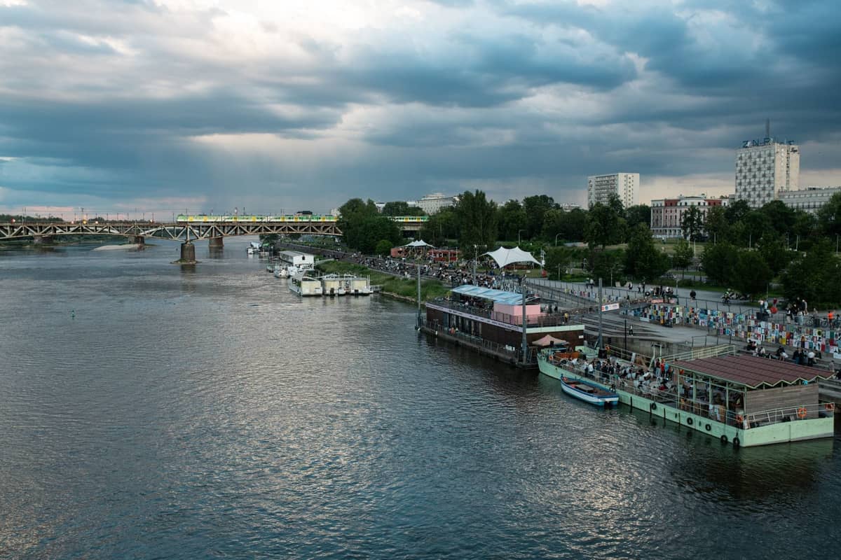Vista del río Vístula con bares flotantes, vegetación ribereña y multitudes disfrutando de la orilla del río en Varsovia, Polonia.
