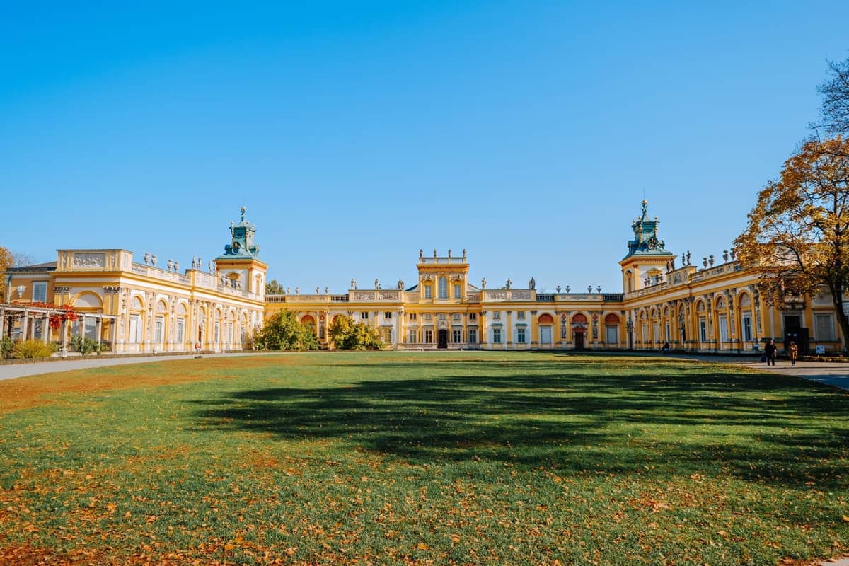 Vista amplia del Palacio de Wilanów con su gran fachada amarilla y un extenso césped verde al frente en un día soleado.
