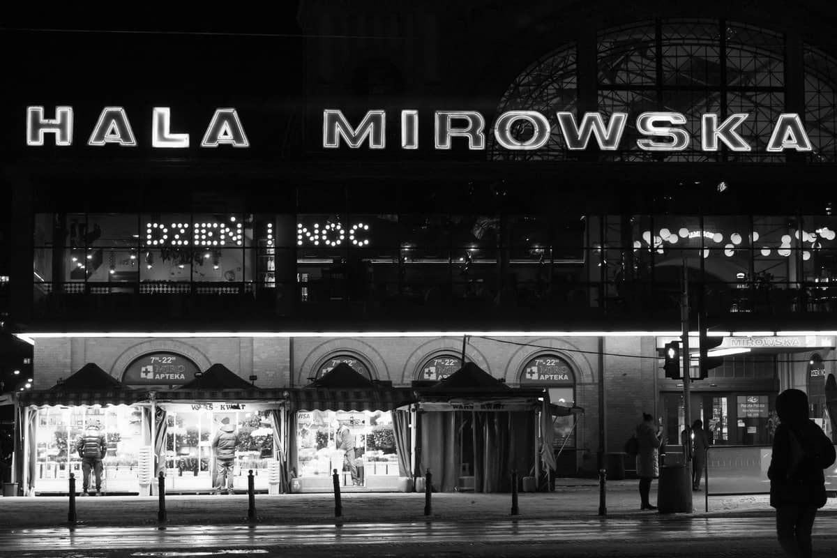 Cảnh đêm của Hala Mirowska ở Warsaw, với các quầy chợ được thắp sáng và biển hiệu lớn phía trên lối vào.