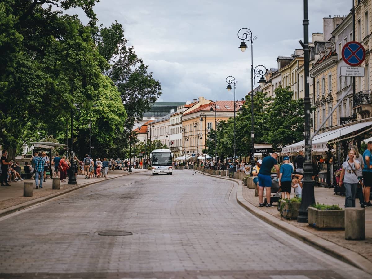 Phố đi bộ rộng rãi và nhộn nhịp ở Warsaw với người đi bộ, cây xanh, mặt tiền lịch sử, quán cà phê và một tàu điện ở phía xa.