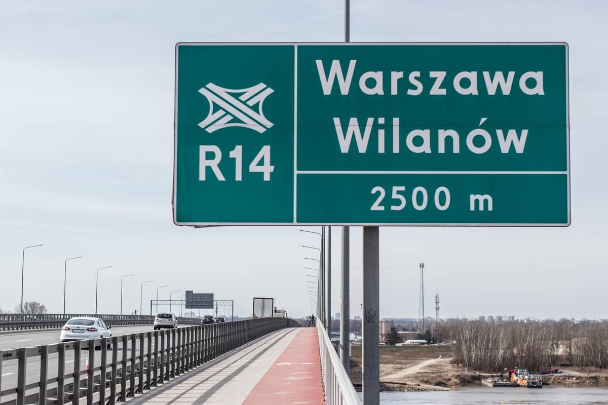 Scena na autostradzie z widocznym znakiem drogowym 'Warszawa Wilanów' i samochodami jadącymi drogą w ciągu dnia.