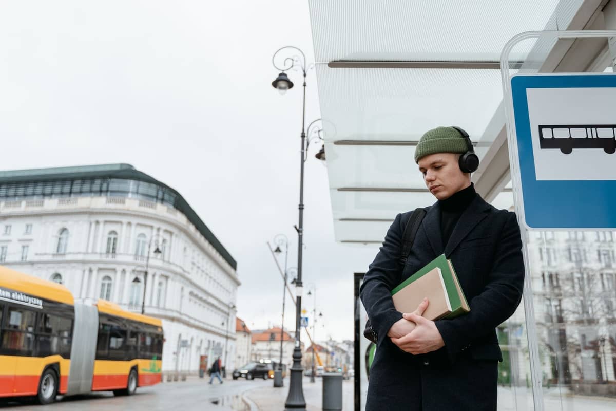 Osoba czekająca na przystanku autobusowym w Warszawie z nadjeżdżającym żółto-czerwonym autobusem, otoczona historycznymi budynkami.