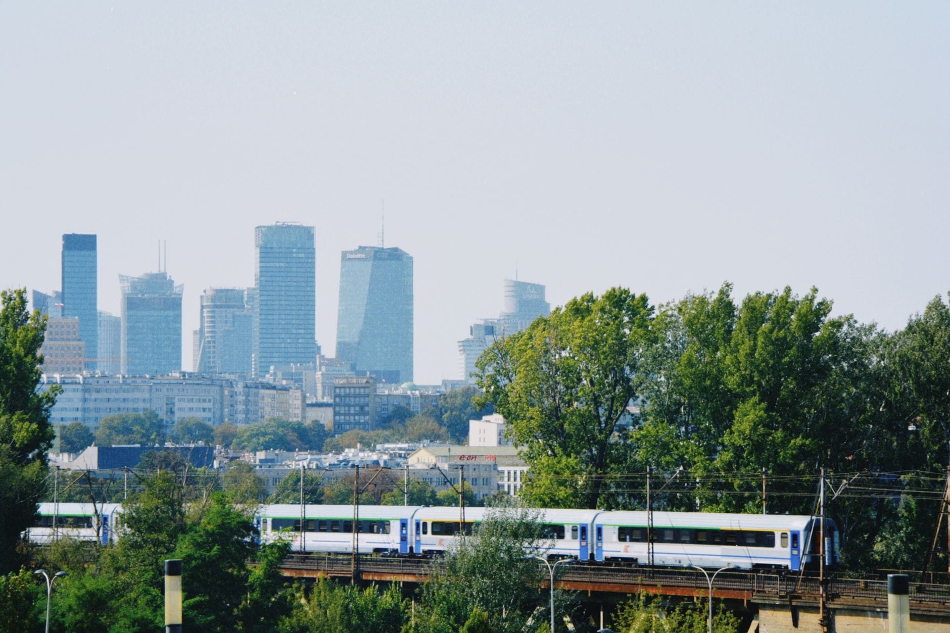 Trem da PKP Intercity passando por um viaduto com o skyline de Varsóvia ao fundo, árvores em primeiro plano e céu limpo.