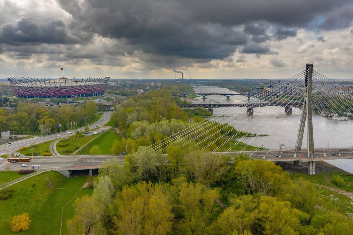 Vista aerea di Varsavia con il fiume Vistola, lo Stadio Nazionale, un ponte strallato e cieli nuvolosi che suggeriscono un tempo variabile.