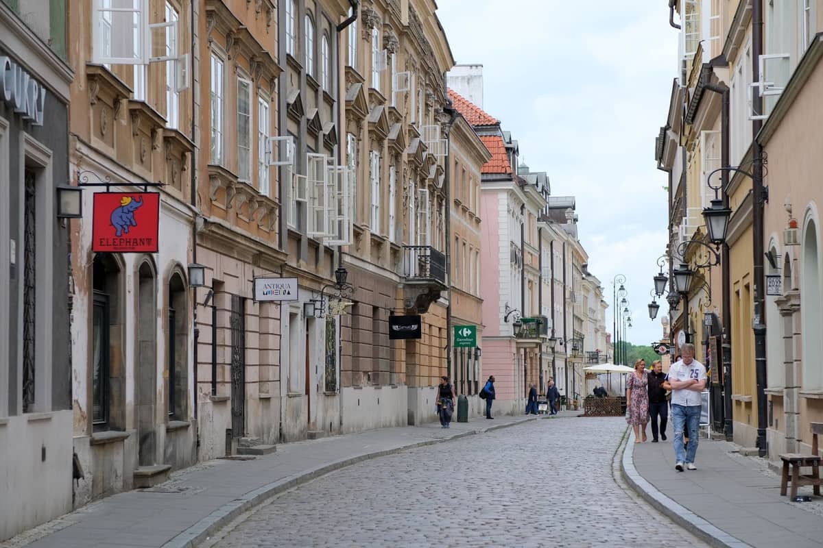 Persone che camminano lungo una strada acciottolata fiancheggiata da edifici storici nel centro di Varsavia in una giornata nuvolosa.