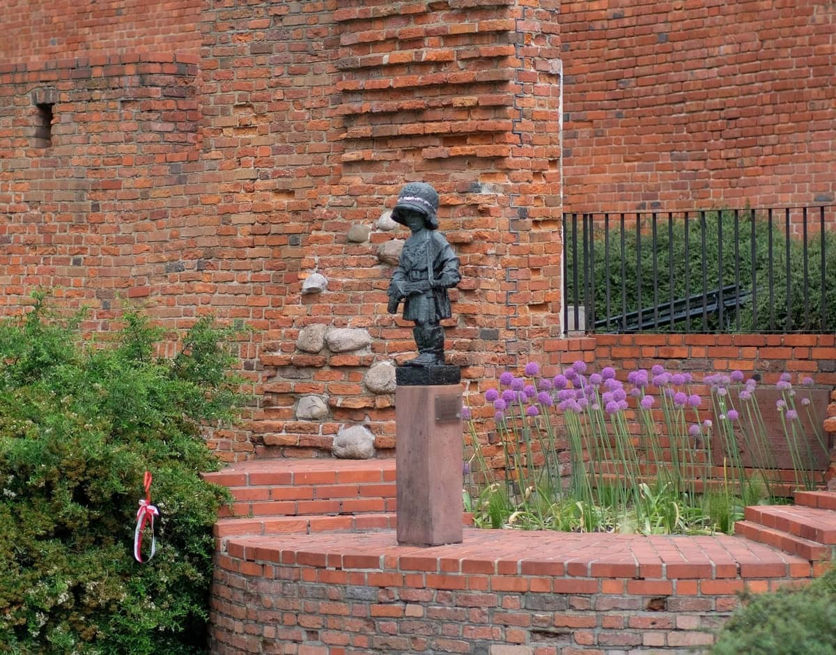 Una piccola statua di un bambino con casco su un piedistallo davanti a vecchi muri di mattoni rossi e cespugli da giardino a Varsavia.