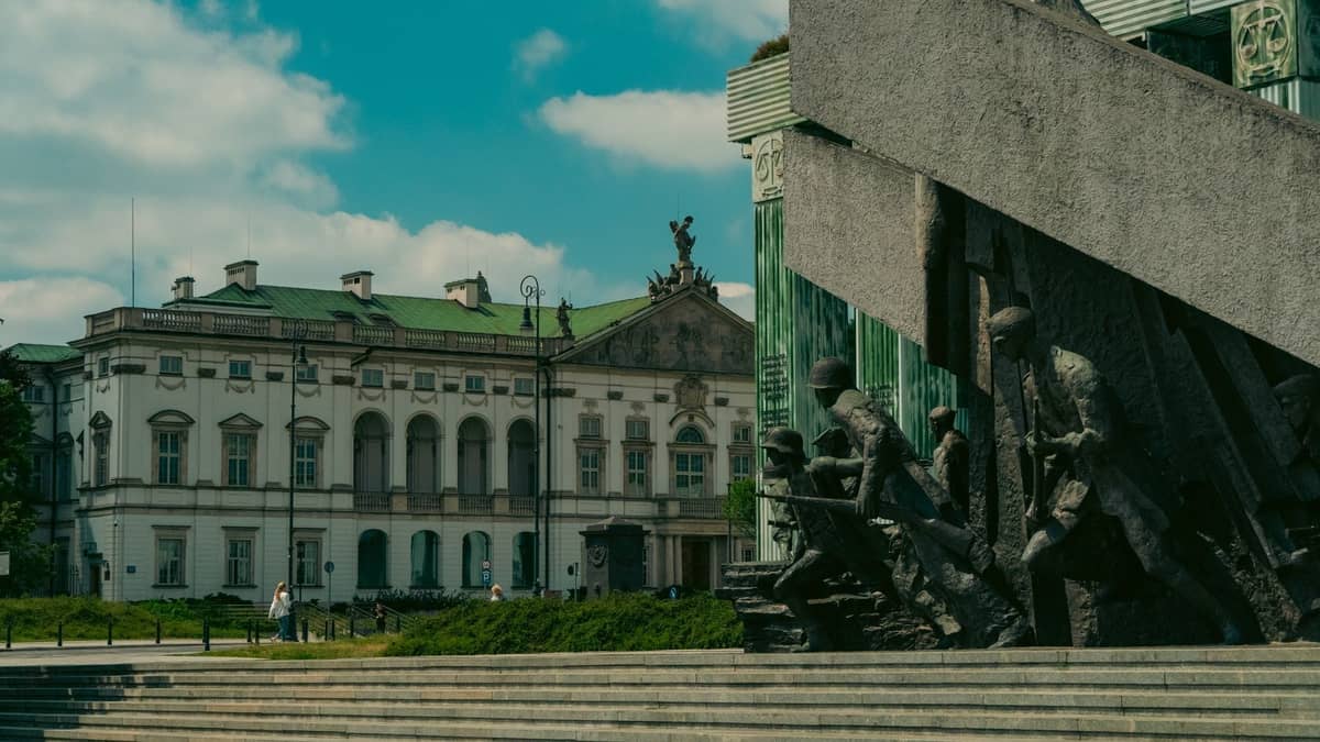Vista ampia del Monumento all'Insurrezione di Varsavia con statue imponenti e un edificio storico sullo sfondo sotto un cielo azzurro.