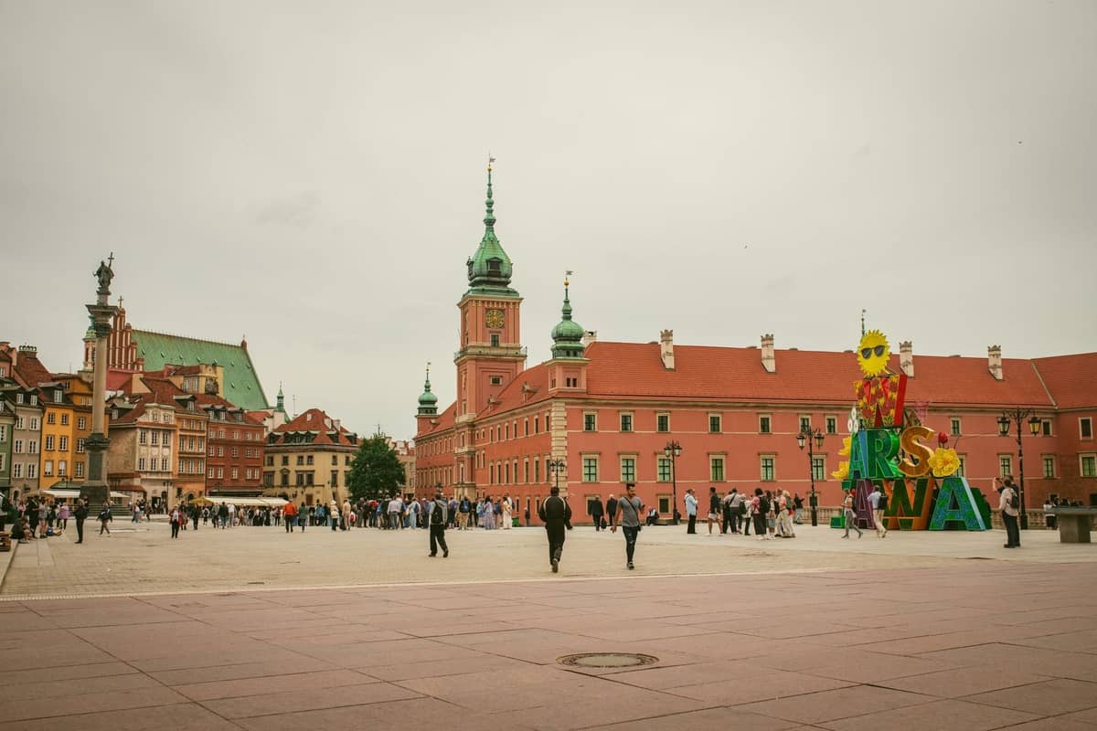 Vista ampia di Piazza del Castello a Varsavia con il Castello Reale, la Colonna di Sigismondo, persone che passeggiano e i colorati edifici della Città Vecchia.