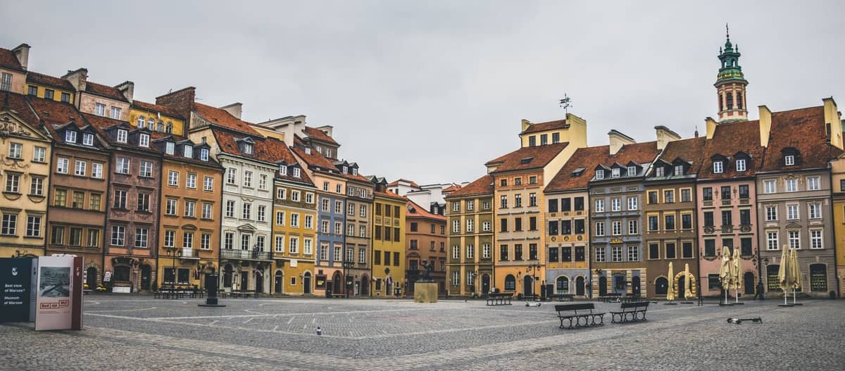 วิวกว้างของจัตุรัสเมืองเก่า Warsaw พร้อมอาคารประวัติศาสตร์หลากสีสัน ยอดโบสถ์ พื้นหินกรวด และม้านั่งใต้ท้องฟ้าที่มีเมฆ