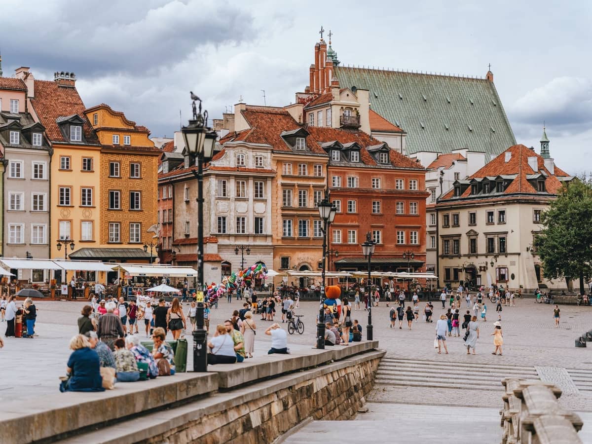 Tłumy ludzi na zrekonstruowanym Rynku Starego Miasta w Warszawie, z kolorowymi fasadami w stylu historycznym i wyraźnym dachem kościoła w tle.