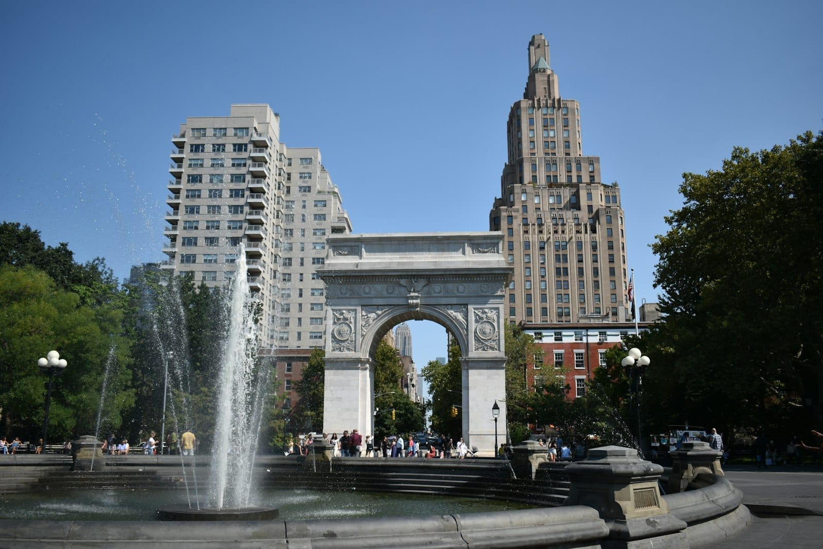 Vista del celebre arco di marmo di Washington Square Park con la fontana centrale, circondato da persone, alberi e gli storici edifici di Greenwich Village su uno sfondo di cielo azzurro.