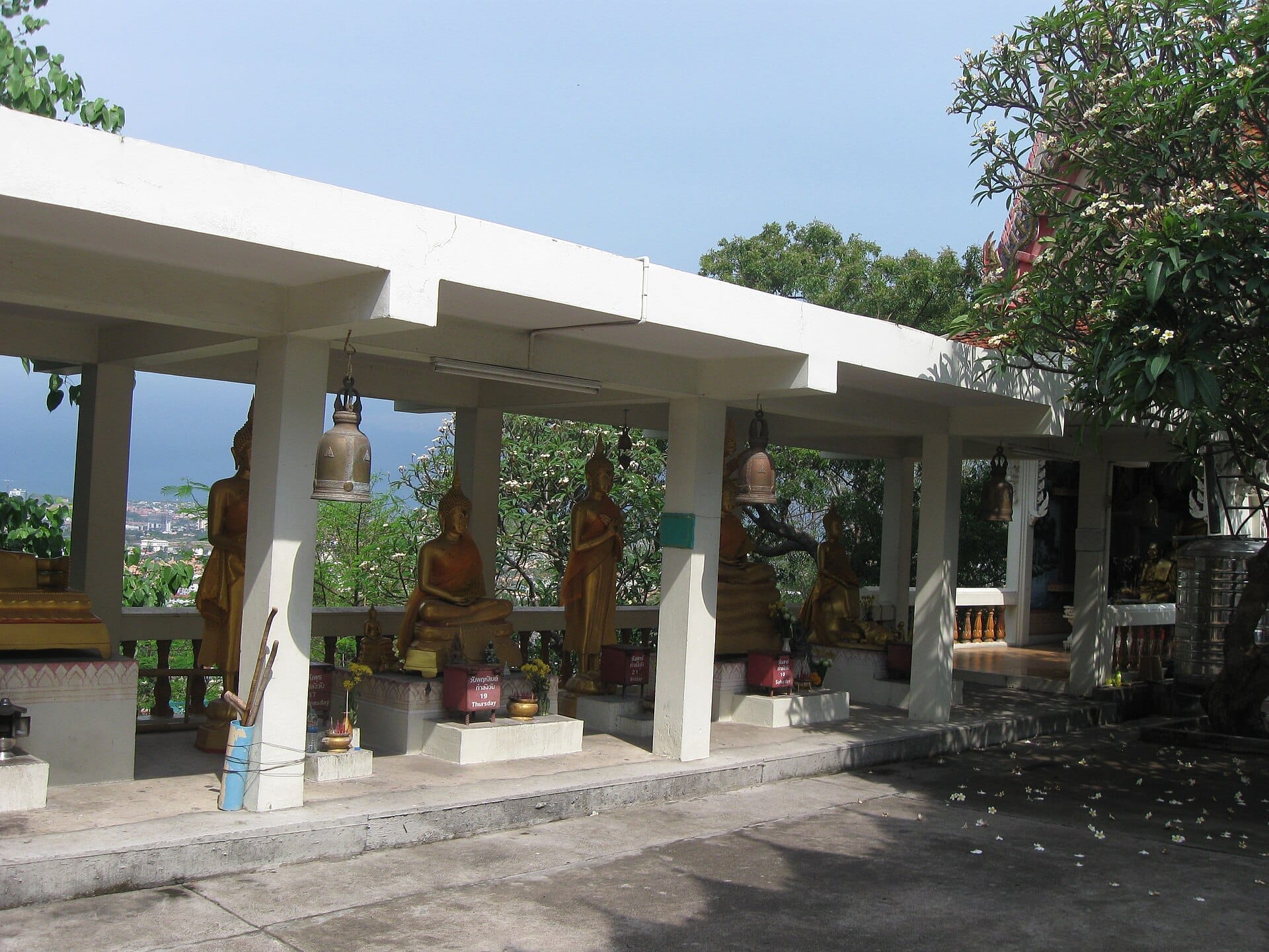 Statue dorate di Buddha sotto un padiglione bianco al Wat Khao Phra Bat, circondate da vegetazione con la baia di Pattaya visibile sullo sfondo.