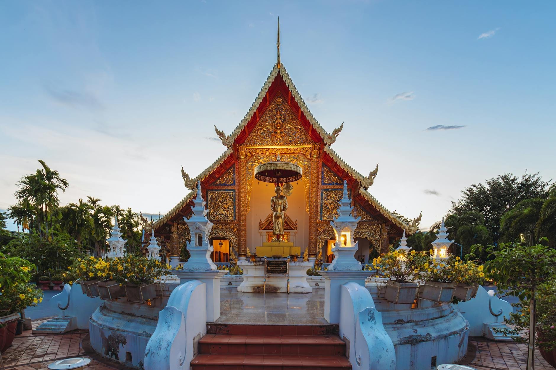 Vista frontal de la sala principal del Wat Phra Singh, ricamente decorada con detalles dorados, rodeada de vegetación y un cielo azul despejado al atardecer.