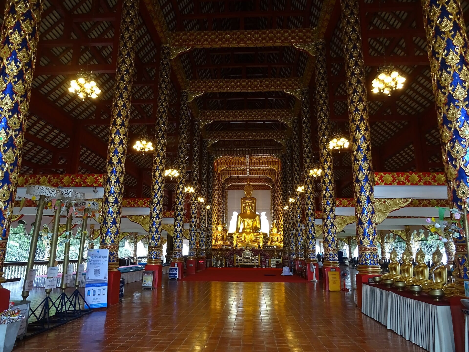 Vista interior del templo Wat Suan Dok, con columnas azules y doradas ricamente decoradas, lámparas de araña y una gran estatua dorada de Buda en el centro.