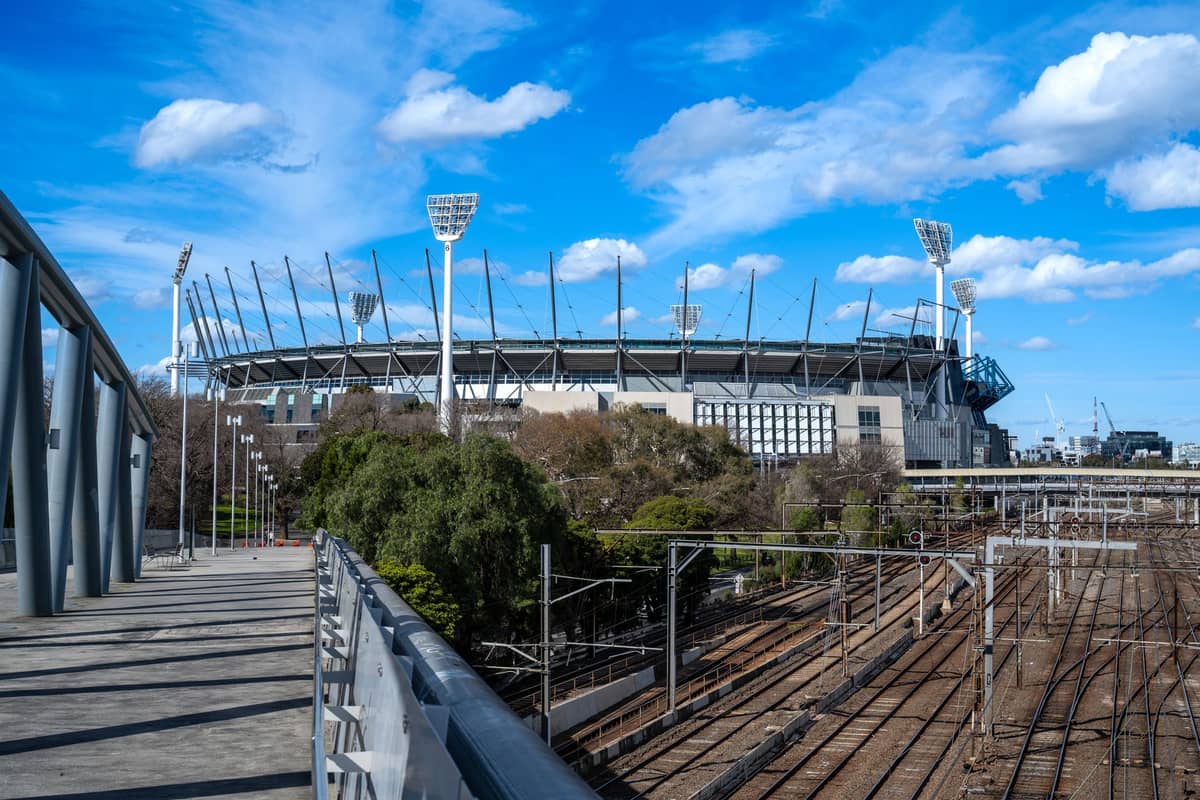Widok zewnętrzny Melbourne Cricket Ground z oświetleniem stadionu i pobliskimi torami kolejowymi pod jasnym błękitnym niebem.