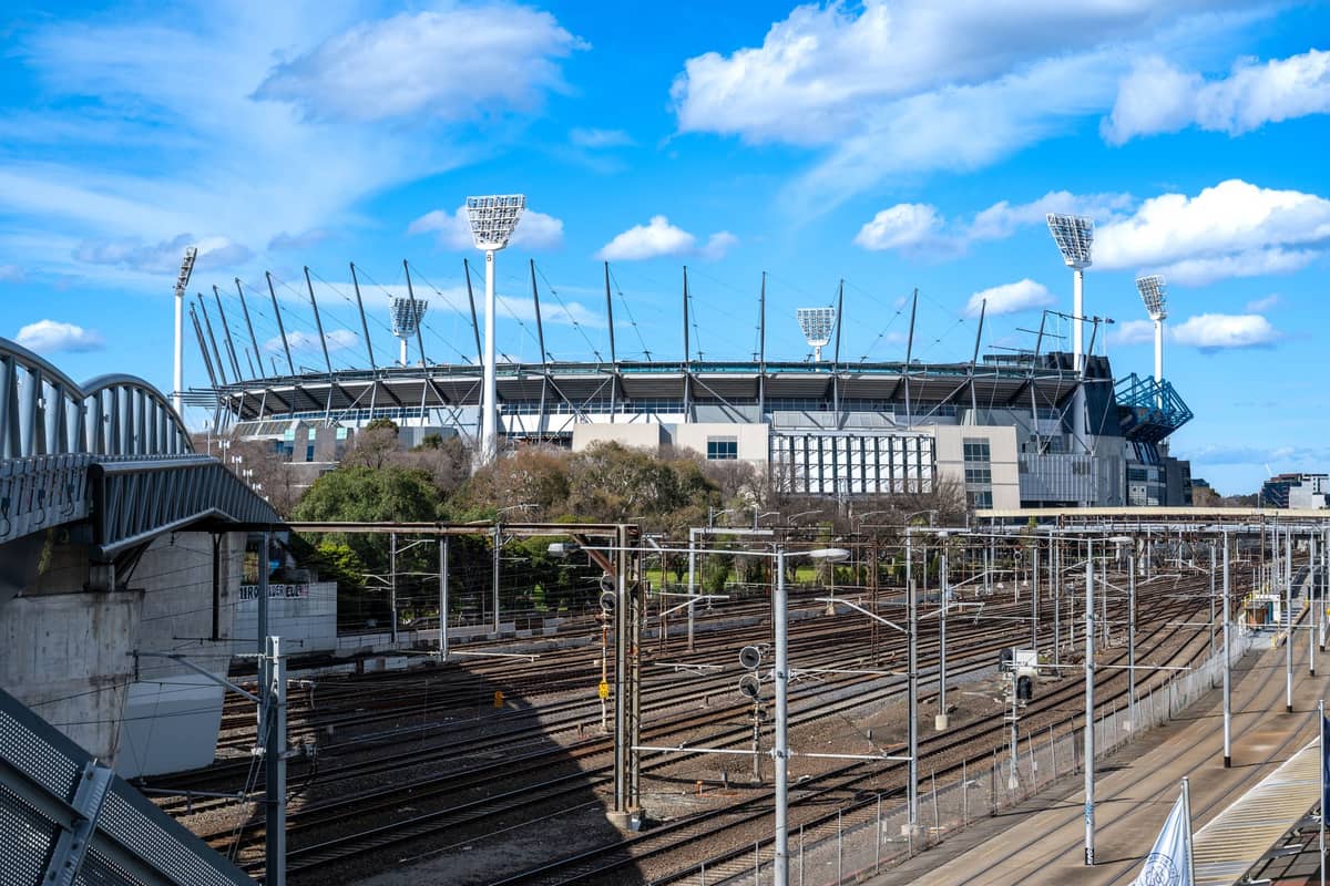 Szeroki widok na stadion Melbourne Cricket Ground (MCG) pod błękitnym niebem, z oświetleniem stadionu i pobliskimi torami kolejowymi.