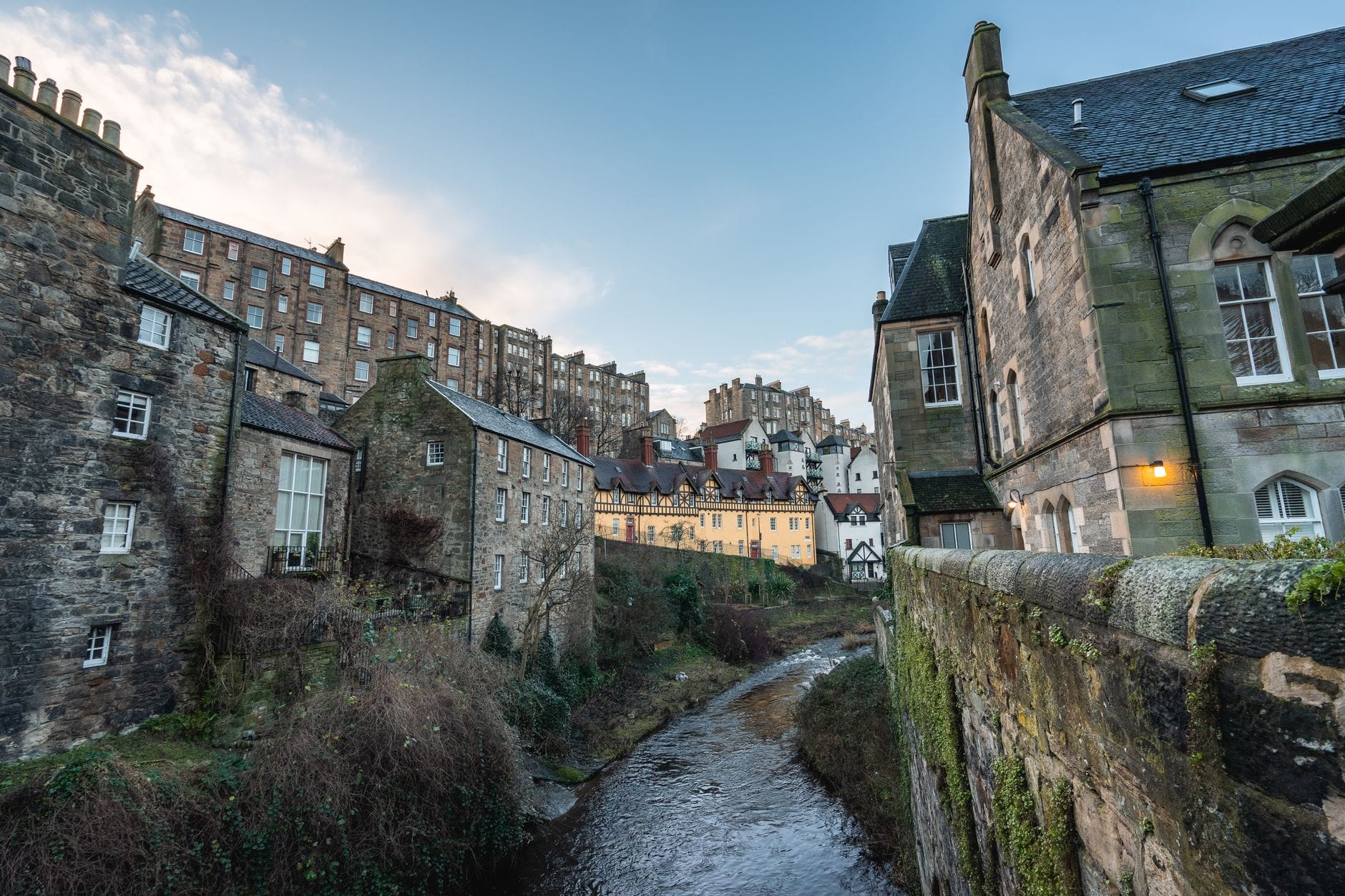 Malerischer Blick entlang des Water of Leith in Dean Village, Edinburgh: alte Steinhäuser am Flussufer, im Hintergrund die Stadt.
