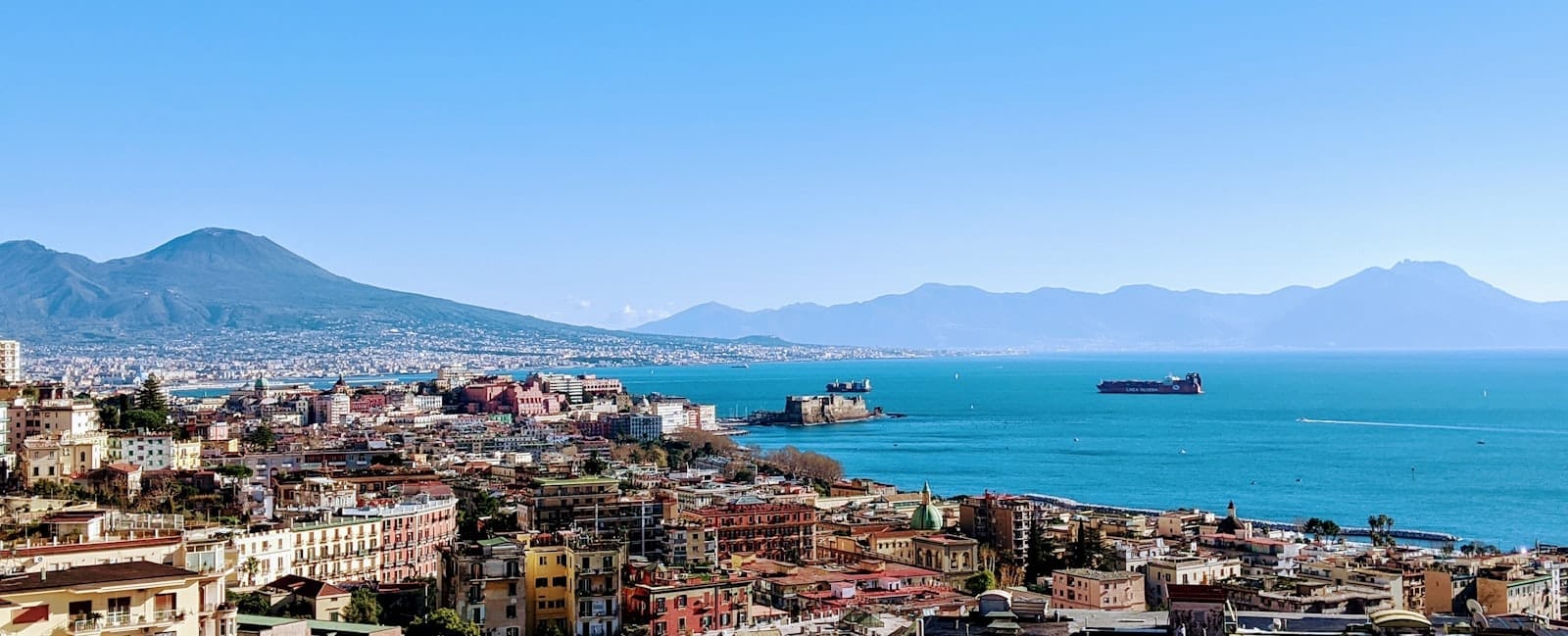 วิวพาโนรามาของริมน้ำเนเปิลส์และลุงโกมาเร พร้อมอ่าวเนเปิลส์ Castel dell'Ovo และภูเขาไฟวิสุเวียสที่มองเห็นได้ในพื้นหลังใต้ท้องฟ้าสีฟ้าใส
