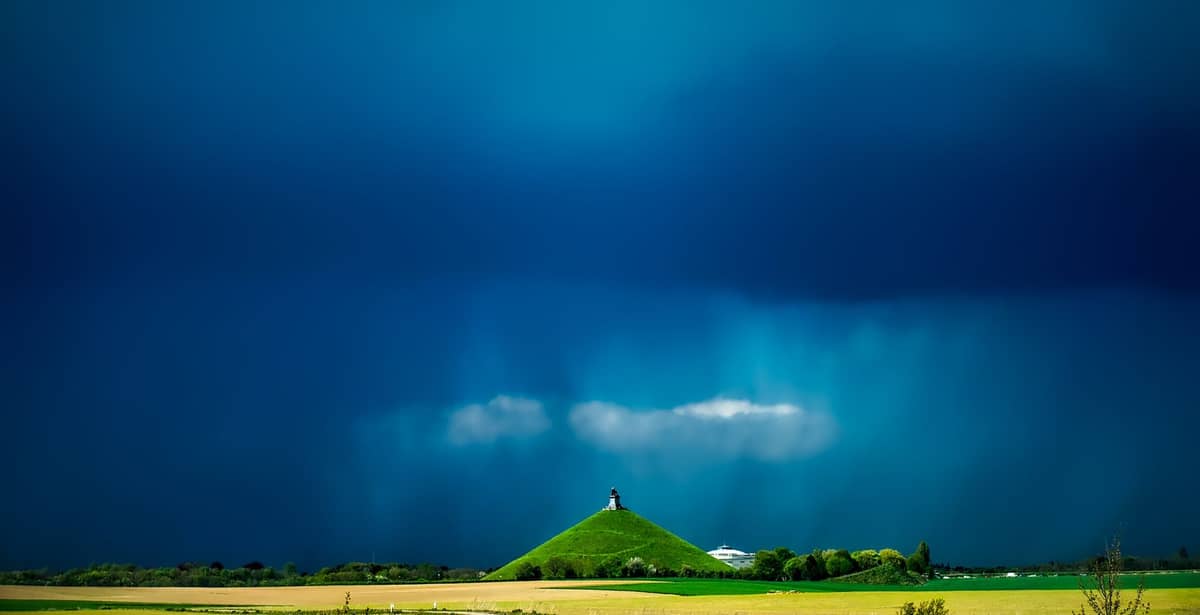 Vista dramática do Monte do Leão se erguendo acima do Campo de Batalha de Waterloo, com a estátua do leão no topo e um céu carregado ao fundo.