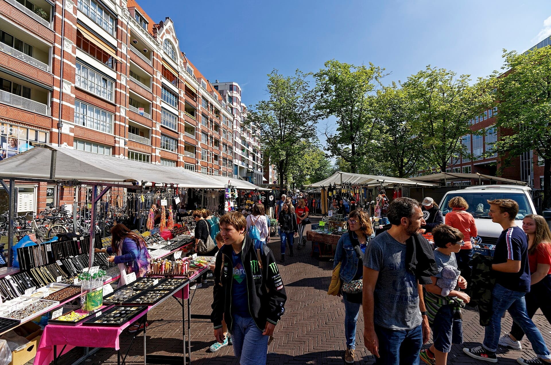 Filas de puestos de mercado y compradores en el Waterlooplein Market de Ámsterdam, con edificios de apartamentos de ladrillo y árboles frondosos bajo un cielo azul brillante.