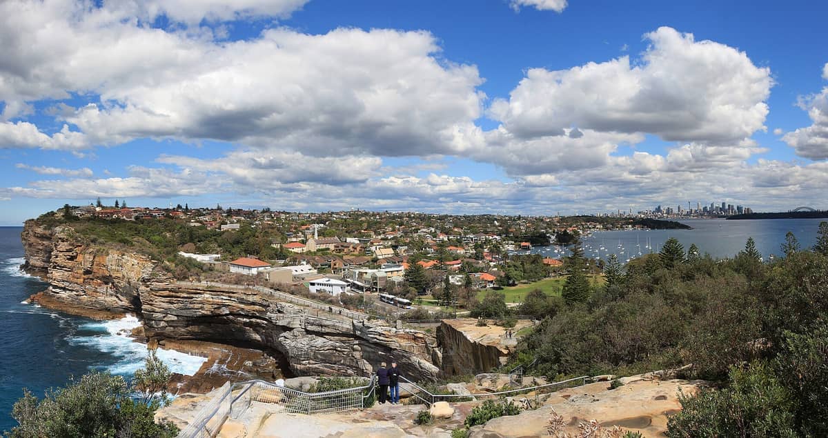 Panoramiczny widok na Watsons Bay z dramatycznymi klifami oceanicznymi, dachami budynków mieszkalnych, spokojnymi wodami portu i panoramą Sydney pod częściowo zachmurzonym błękitnym niebem.
