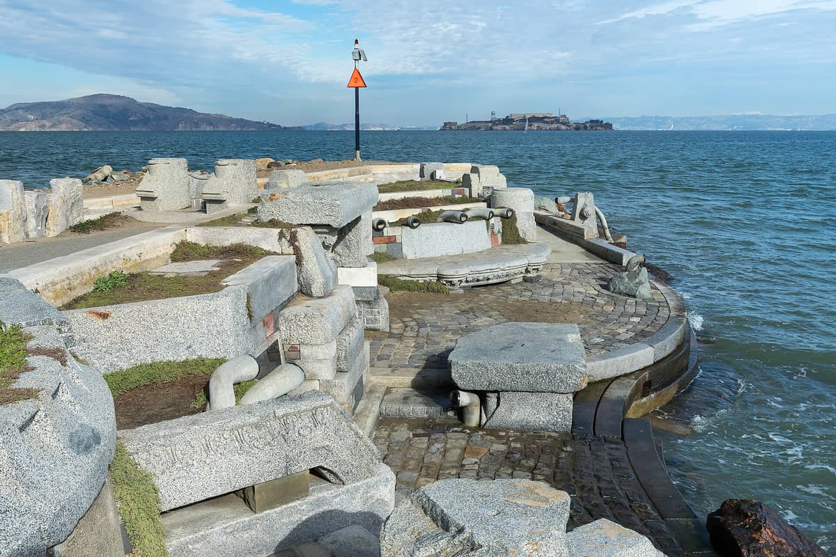 Vista amplia del Wave Organ en San Francisco con bancos de granito esculpido, tubos y trabajo de piedra sobre un espigón rodeado de agua de la bahía y la isla de Alcatraz al fondo.