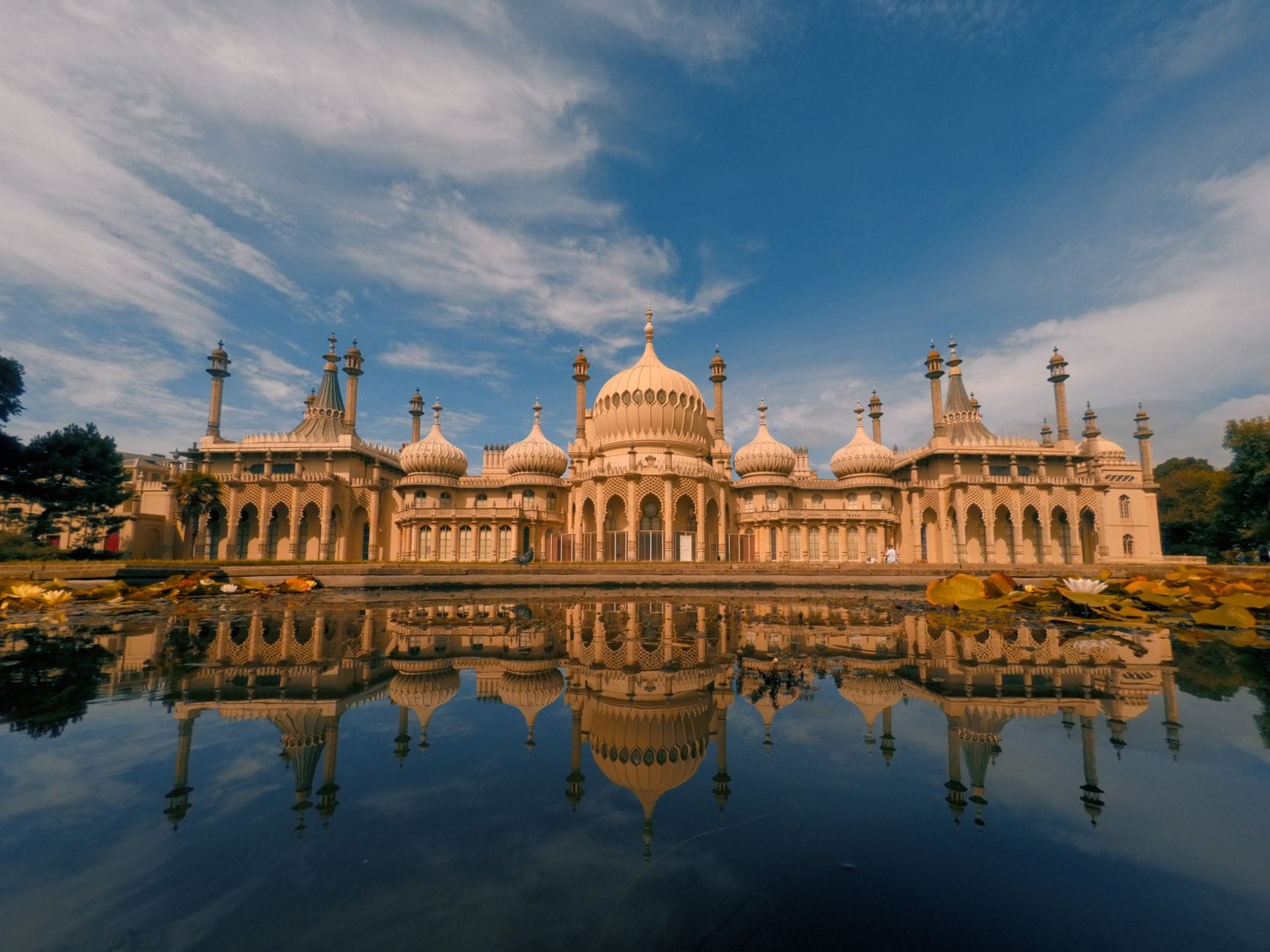 Vista ampia del Royal Pavilion a Brighton con le sue cupole ornate e il riflesso in uno stagno sotto un cielo drammatico.
