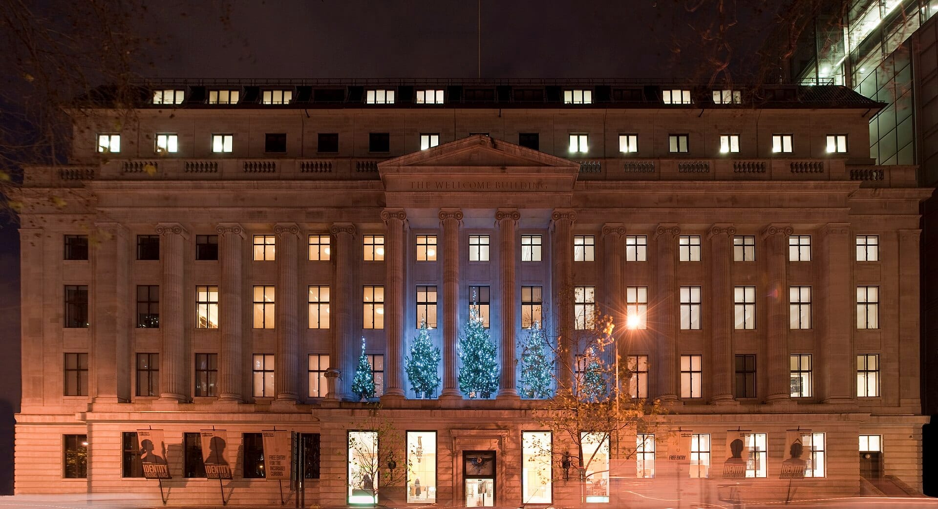 Vista frontale della Wellcome Collection di notte, con la sua grandiosa facciata neoclassica illuminata da luci calde e decorata con alberi festivi illuminati di blu.