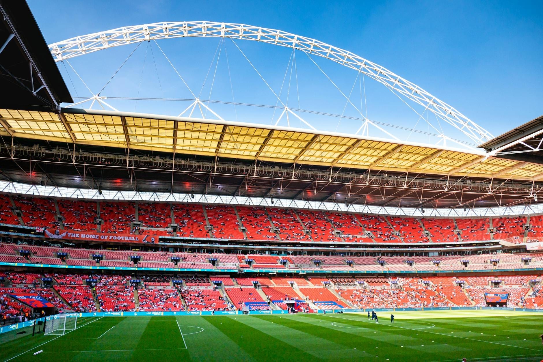 Vista grandangolare dell'interno del Wembley Stadium con il suo iconico arco bianco, il manto erboso verde e i sedili rossi sotto un cielo azzurro limpido.