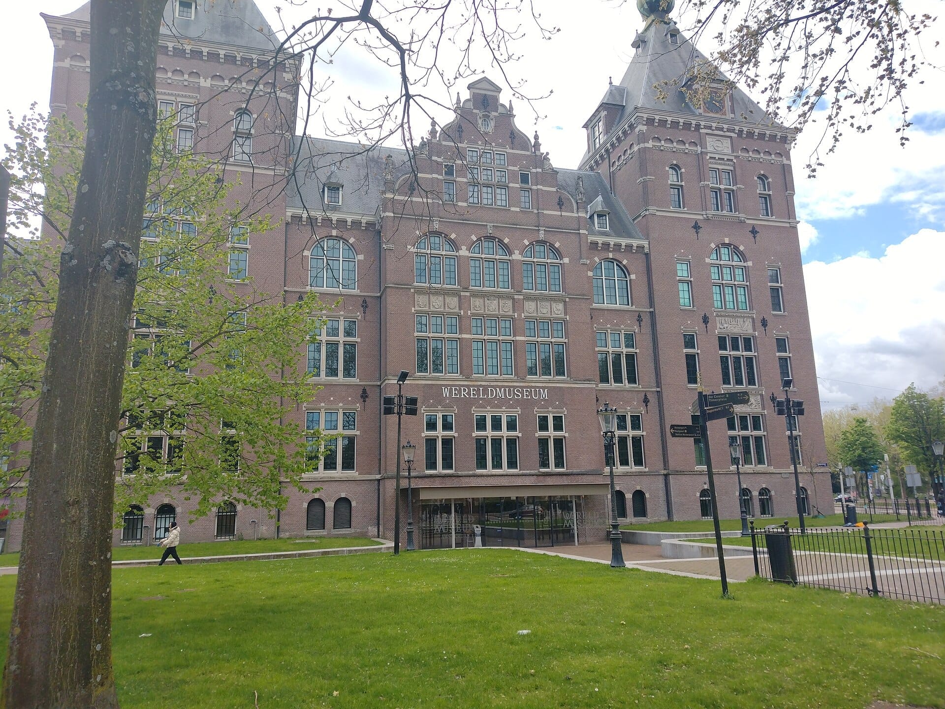 La fachada del Wereldmuseum Amsterdam, un gran edificio histórico de ladrillo con grandes ventanas, jardines verdes y árboles alrededor en un día nublado.