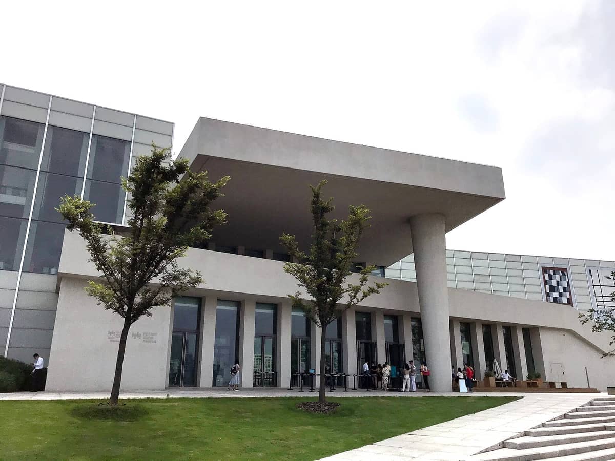 Moderne Fassade des West Bund Museum in Xuhui, Shanghai, mit minimalistischer Architektur, geometrischen Formen, kleinen Bäumen und Besuchern auf dem Rasen.