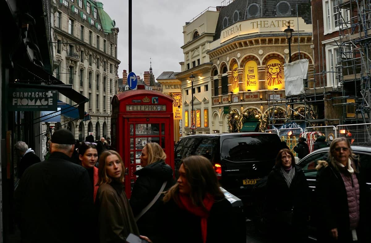 Rua movimentada no West End de Londres com o Lyceum Theatre, uma cabine telefónica vermelha e pedestres, destacando um clássico marco teatral e uma atmosfera vibrante.