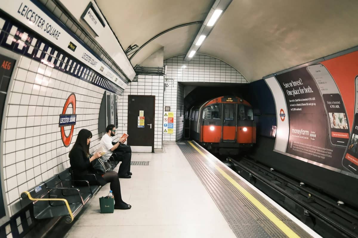 Plataforma do metrô de Londres na estação Leicester Square com pessoas esperando e um trem chegando, paredes de azulejos e sinalização clara visíveis.