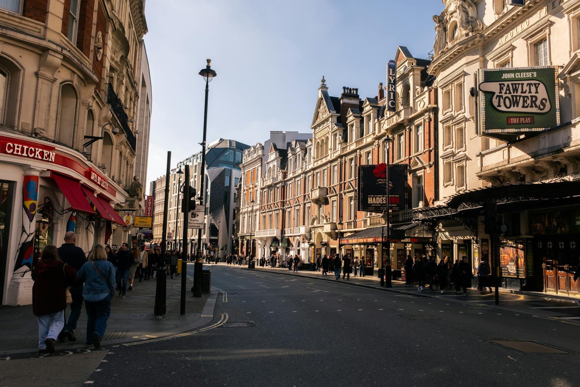 Vista ampla de uma rua do West End de Londres sob céu azul, com teatros históricos exibindo cartazes de espetáculos, cheios de pessoas e arquitetura clássica, transmitindo a atmosfera vibrante do bairro teatral.
