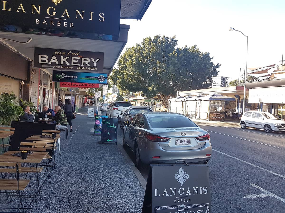 ภาพถนน Boundary Street ในย่าน West End ผู้คนจิบกาแฟริมฟุตบาท ร้านท้องถิ่น และรถจอดเรียงรายในวันที่แดดสดใส