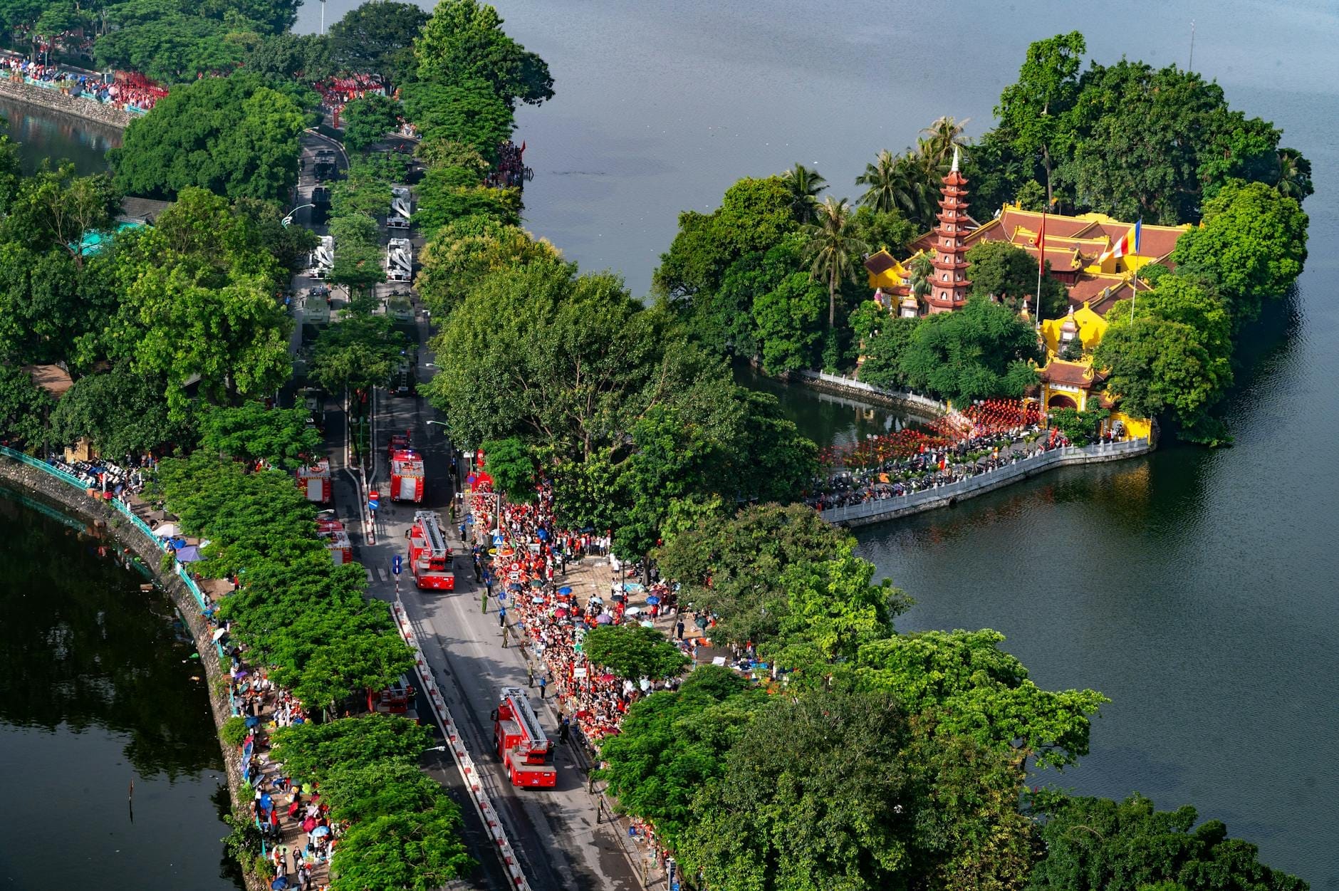 Aerial view of West Lake in Hanoi with the Tran Quoc Pagoda surrounded by lush trees, crowds, and fire trucks along the lakeside road.