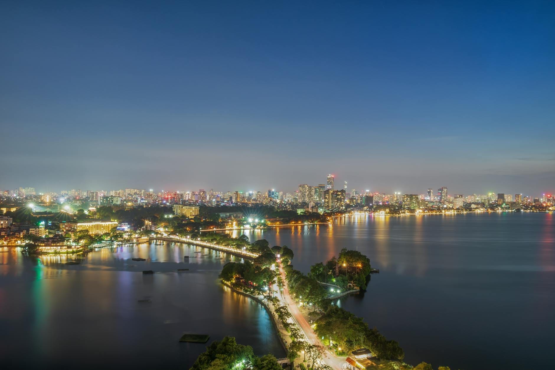 A stunning aerial sunset view over West Lake in Hanoi, with a curving tree-lined boulevard, city lights, and calm waters reflecting the vibrant evening sky.