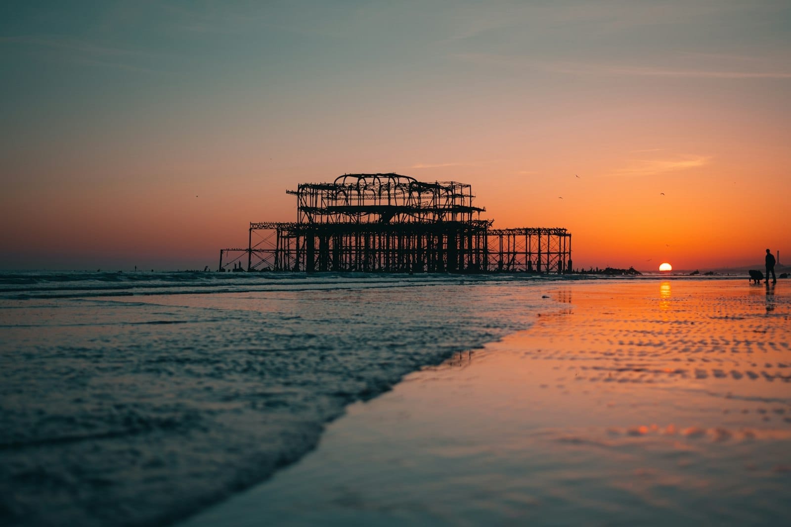 The skeletal remains of West Pier Ruins stand offshore in Brighton at sunset, reflected in the wet sand and calm sea.