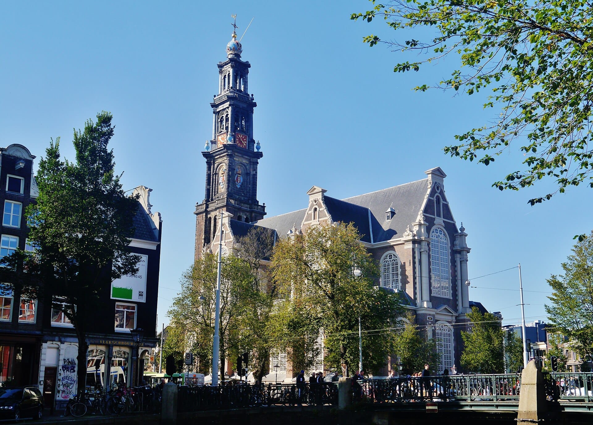 La iglesia Westerkerk en Ámsterdam se alza majestuosa junto al canal Prinsengracht, rodeada de árboles y clásicos edificios holandeses en un día soleado.
