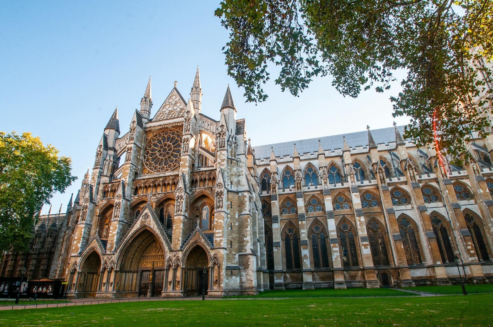 Vista frontale dell'imponente facciata gotica di Westminster Abbey con il rosone, incorniciata da prato verde e alberi in una luminosa giornata di sole.